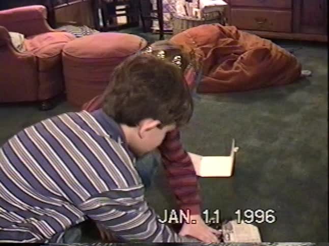 A young boy wearing a birthday crown opens a card and a gift from his brother in their living room in 1996