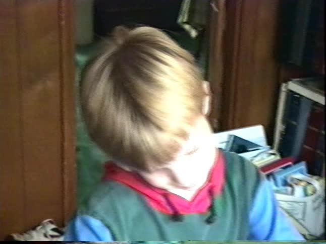 Young boy in a colorful hoodie sits at a wooden table indoors, concentrating on a small craft project