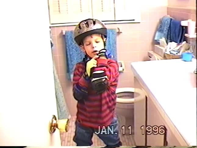 A young boy proudly shows off his full set of rollerblading gear in a bathroom in a 1996 home video