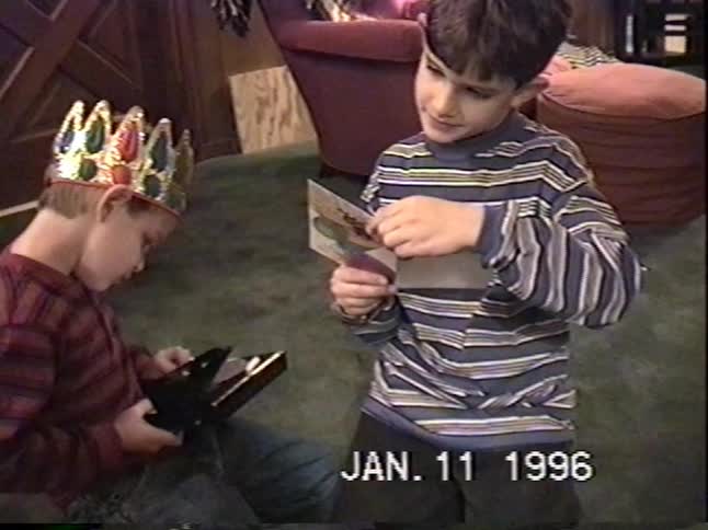 Two young brothers celebrate a birthday in 1996, with one wearing a crown while they open a gift and read a card together