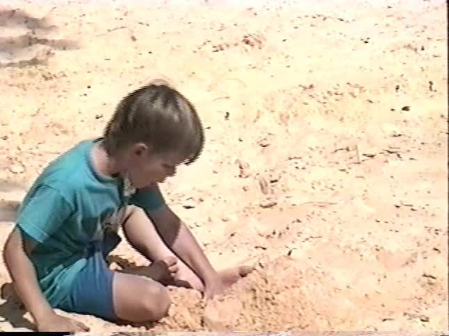 A young boy in a blue shirt and shorts plays alone in a sand volleyball court in a park on a sunny day