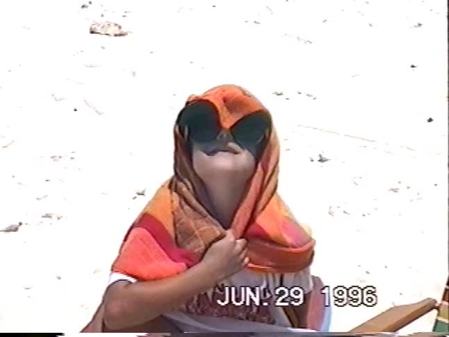 A quirky girl with a towel over her head and oversized sunglasses sits on a sandy beach on a sunny day in 1996