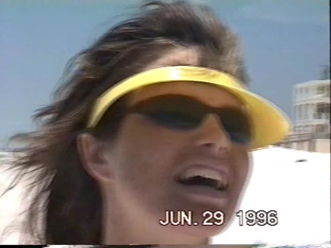 A woman in a yellow visor smiles on a windy beach, followed by a shot of a young boy wrapped in a towel in a 90s home video