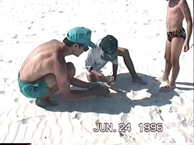 A father and his two young sons light a small firework in the white sand on a sunny day at the beach in 1996
