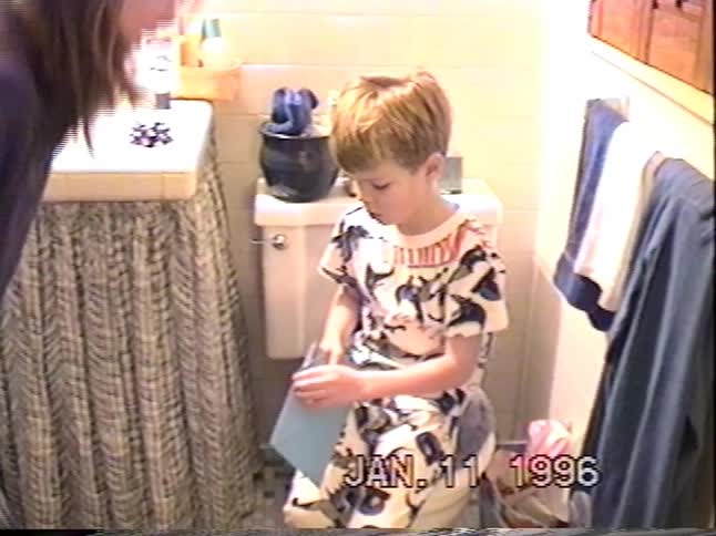 Young boy in patterned pajamas opens a card with help from a woman in a small bathroom in vintage home video footage