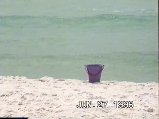 A woman in a swimsuit smiles and holds a boogie board on a white sand beach before heading into the water in this vintage 1996 home video
