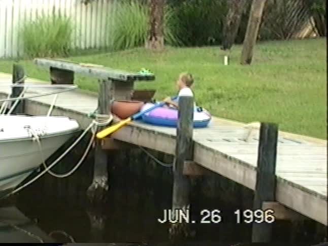 A young boy happily plays in an inflatable raft on a wooden dock before two older boys are seen fishing in the marina on a summer day in 1996