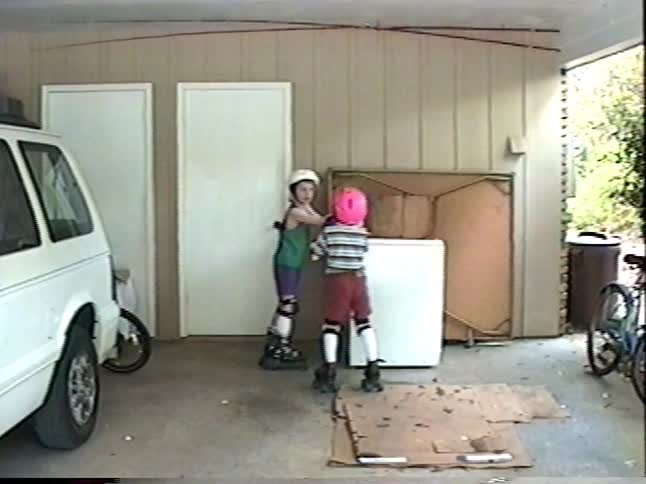 A young boy gets help from his sibling with his helmet before they practice roller skating together in a vintage home video