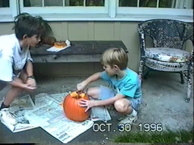 Two young brothers work together to carve a jack-o'-lantern on their porch in a nostalgic home movie from 1996