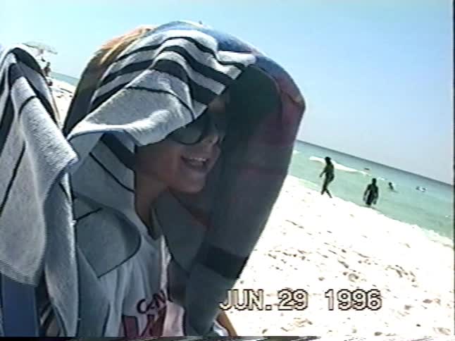 A vintage home video from 1996 shows a man wading in the ocean and a child relaxing on a sunny, white sand beach