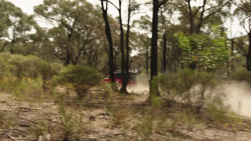 A red four-wheel-drive truck with a rooftop tent kicks up a cloud of dust while driving on a dirt track through a forest