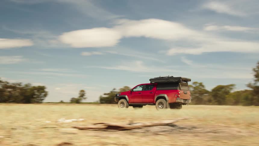 A red overland truck kicks up dust as it drives off-road through a dry, grassy field in the Australian outback on a sunny day