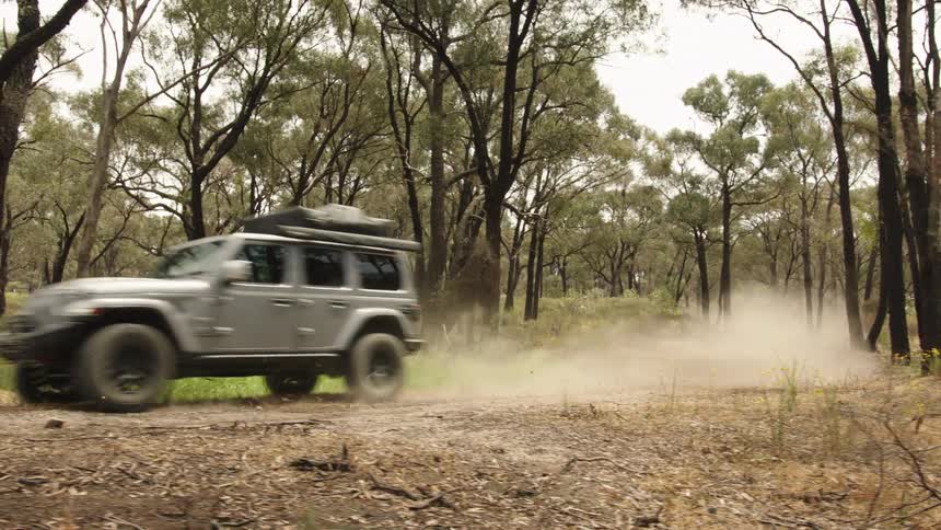 A silver Jeep Wrangler kicks up a large cloud of dust as it drives in slow motion along a dirt track through a forest
