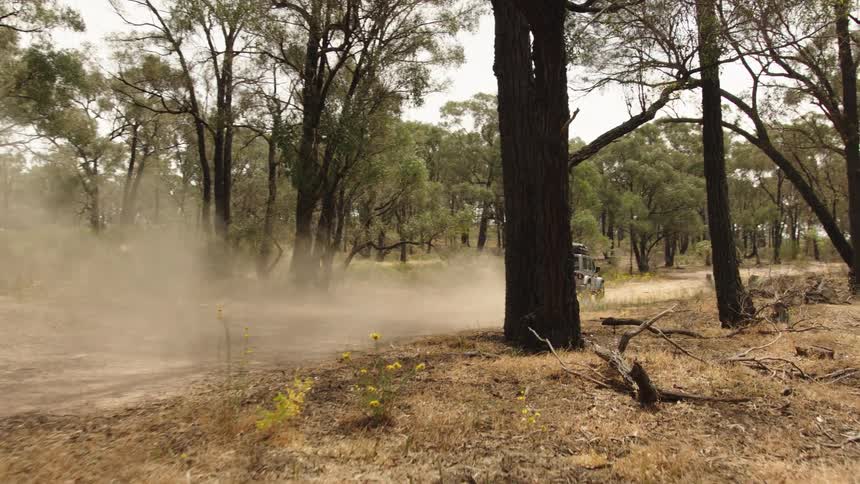 An SUV with a rooftop tent drives down a dusty dirt road through a dry forest, kicking up dust in its wake
