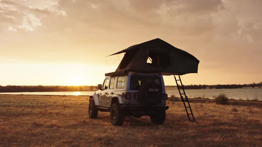 A Jeep Wrangler with a rooftop tent is parked in a golden field next to a lake at sunset