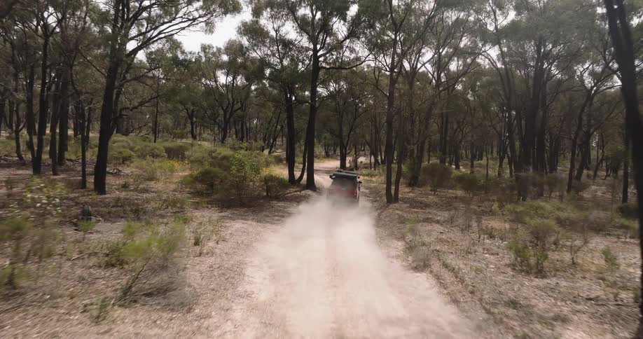 Aerial shot follows a red off-road truck with a rooftop tent as it drives away on a dusty trail through the Australian bush
