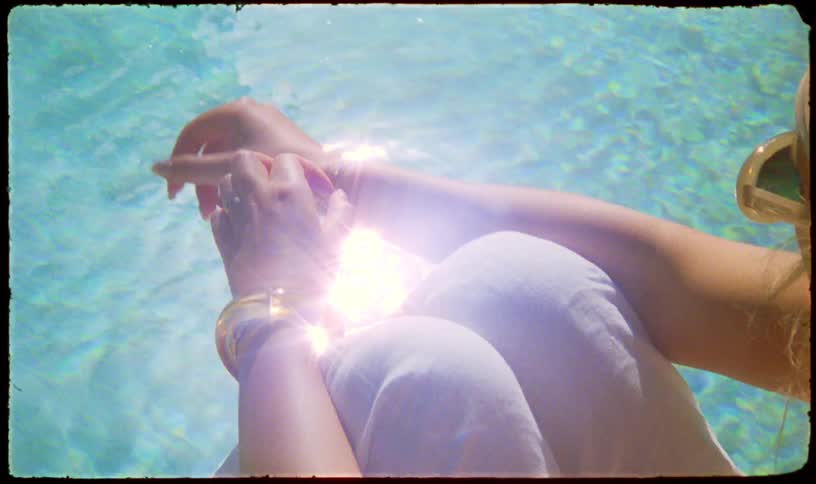 A vintage-style shot shows a woman relaxing by a sparkling swimming pool, her hands clasped as a bright sun flare shines