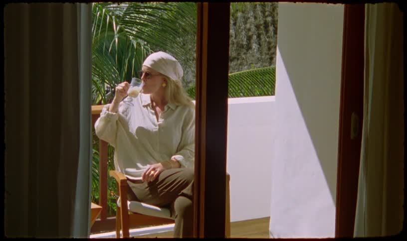 A woman in a headscarf and sunglasses relaxes on a sunny tropical balcony, sipping a drink and enjoying her vacation