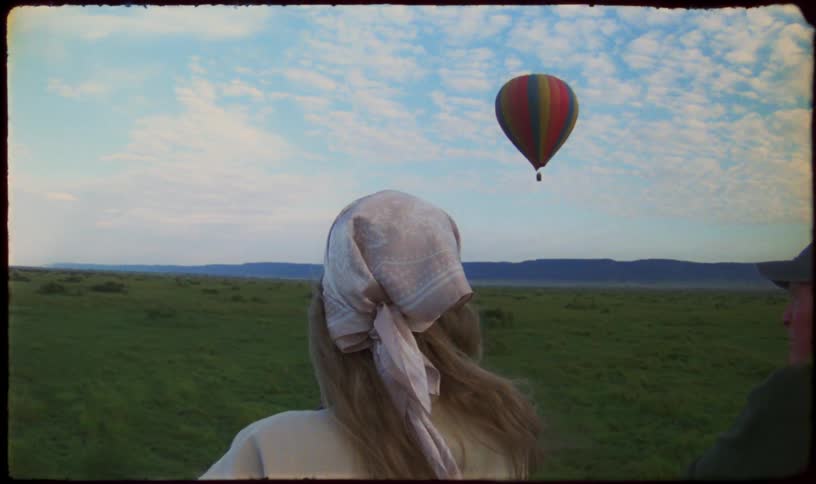 A woman in a hot air balloon watches another balloon float gracefully over the vast, green plains