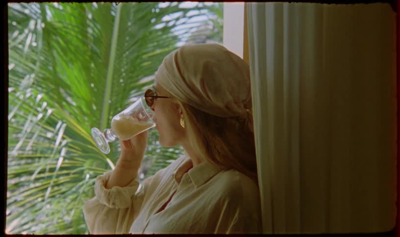 A stylish woman in a headscarf relaxes by a window, sipping a tropical drink and looking out at palm trees on a sunny day