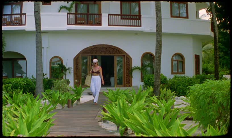Vintage footage shows a stylish woman walking out of a luxurious tropical resort onto a wooden path surrounded by lush greenery
