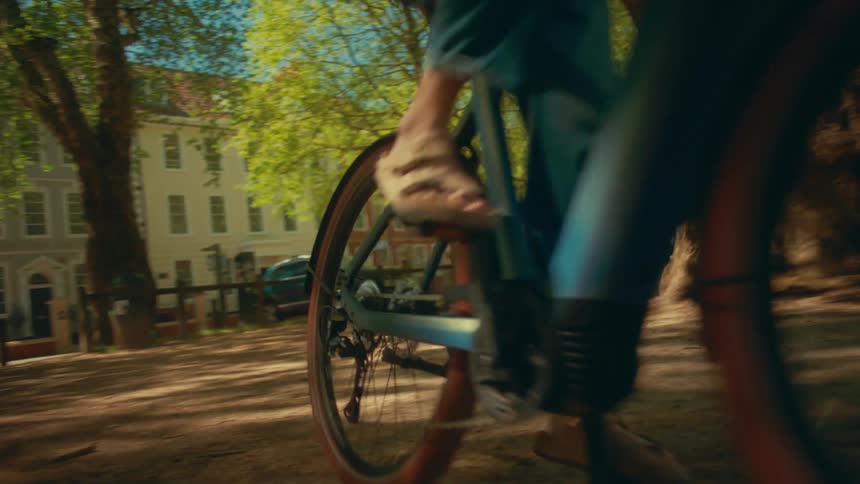 A woman in a helmet rides her bicycle through a sunny city park with trees and historic buildings in the background