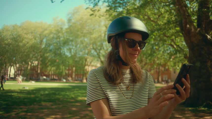 A smiling young woman in a bicycle helmet and sunglasses enjoys a sunny day in the park while scrolling on her smartphone