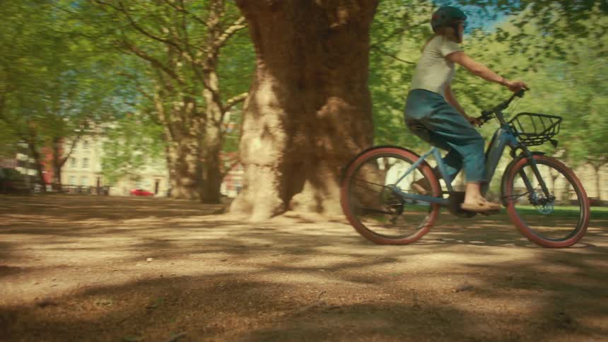 A woman on a bicycle rides along a dirt path in a city park under the dappled sunlight of large trees