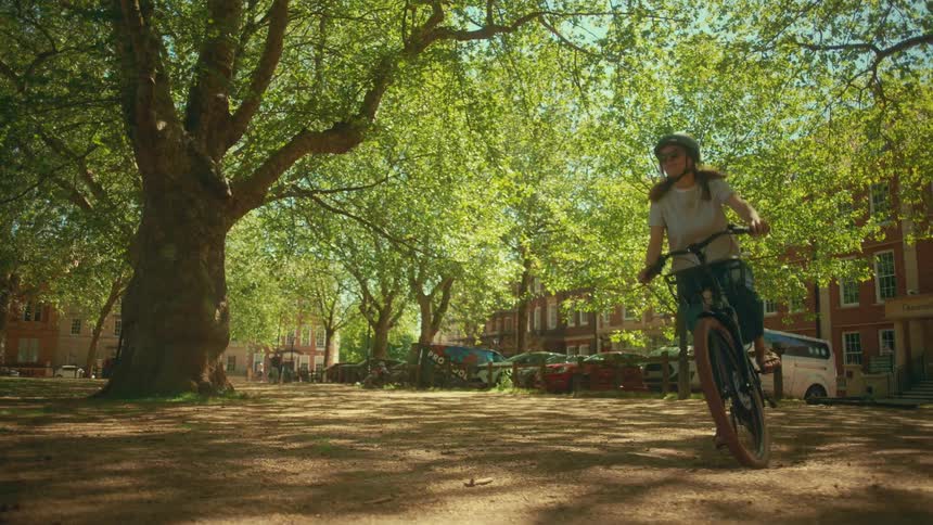 A woman in a helmet rides her bike through a sunlit city park with lush green trees in slow motion