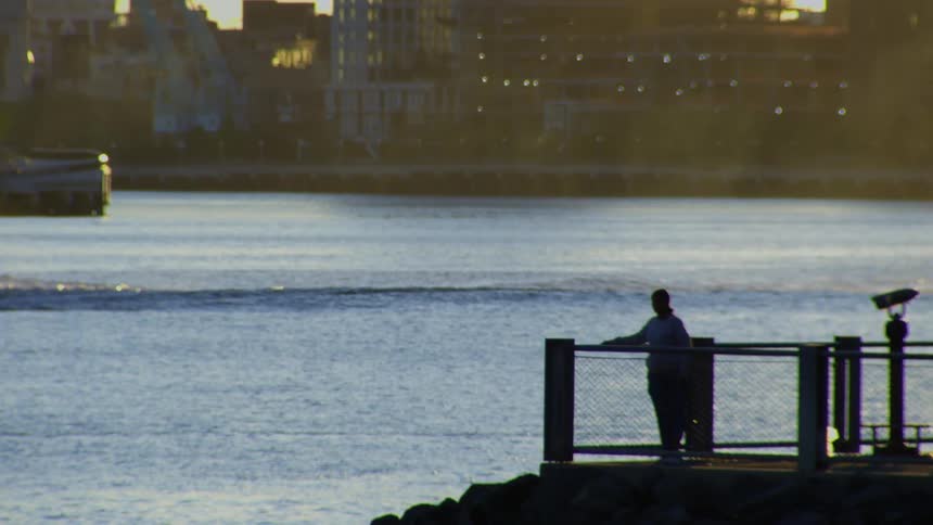 A silhouetted person stands alone on a pier at sunset, looking out at the shimmering water with the city skyline in the background