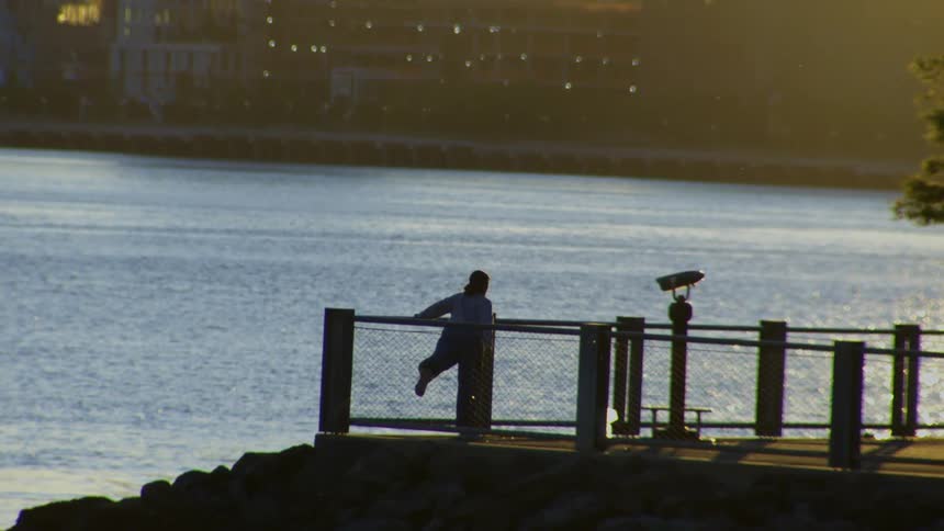 A silhouetted person stands alone on a pier at golden hour, contemplating the tranquil water and distant city lights