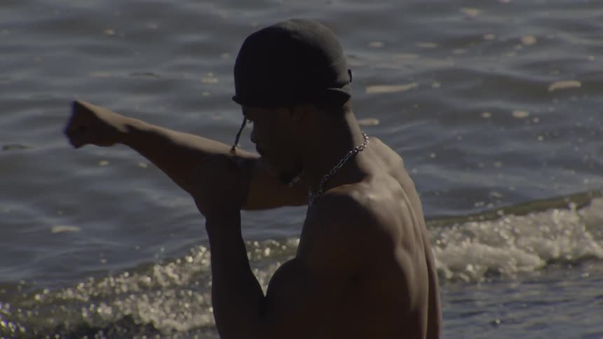 Slow motion footage from behind of a muscular Black man shadow boxing in front of the water