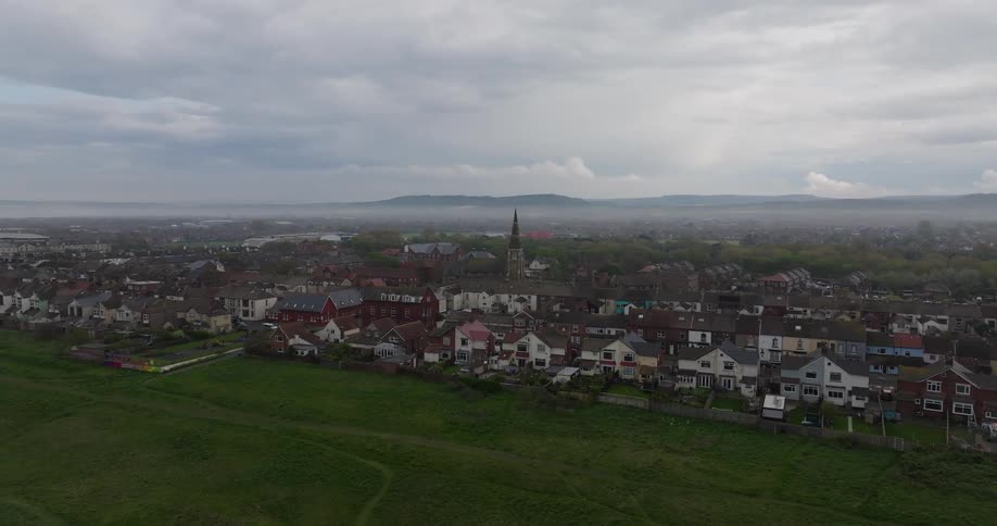 A slow aerial push-in shot reveals a quaint English town with a central church steeple, nestled by a green hill on a cloudy day