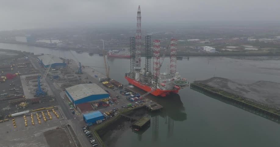 Aerial view of a massive jack-up rig docked in an industrial port on a foggy and overcast day