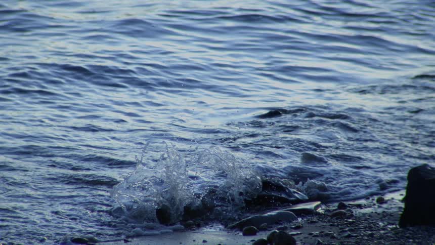 A tranquil wave splashes against rocks on the shoreline in mesmerizing slow motion