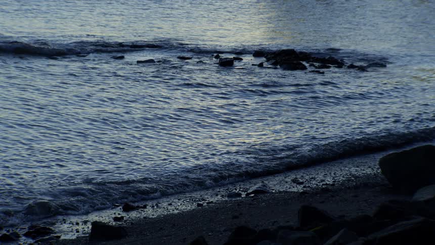 The tranquil blue waters of the ocean gently roll onto a rocky shoreline in slow motion