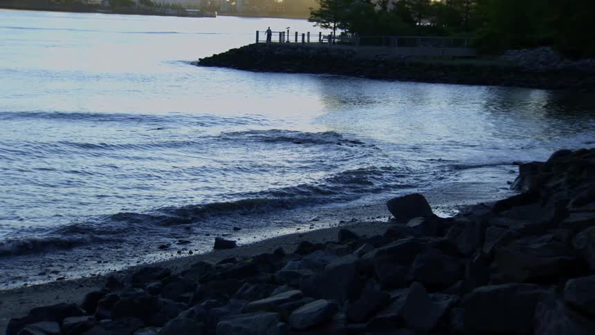 Gentle waves wash onto a rocky riverbank at twilight as people stand on a distant pier