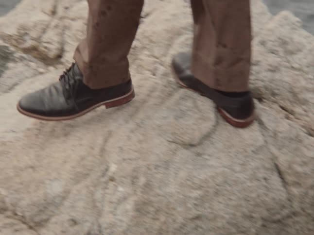 A man in brown pants and black dress shoes carefully walks across rocks next to the water