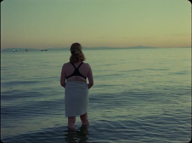 A woman wearing a bikini top and a towel stands in the ocean at dusk, looking out at the horizon