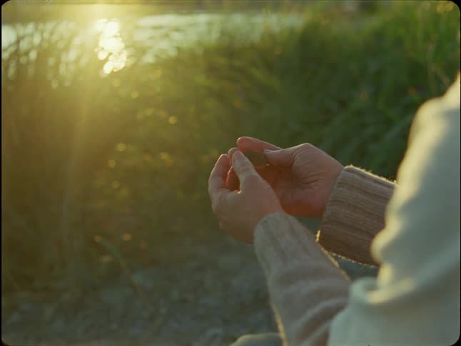 A close up of a person's hands as they gently close around a small rock while sitting in nature during golden hour