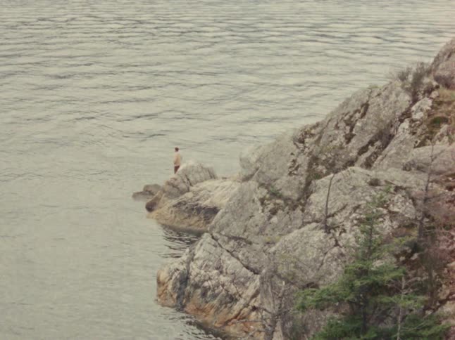 Vintage footage shows a lone fisherman casting his line from a rocky outcrop into a vast, calm lake