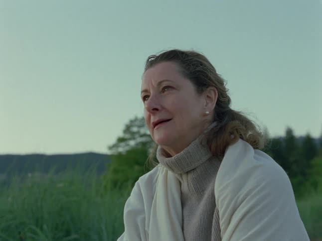 A pensive older woman wrapped in a blanket smiles fondly as she reflects while sitting outdoors at dusk