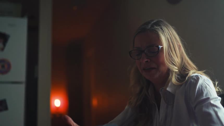 A distraught blonde woman with glasses cries and argues passionately in a dimly lit room at night