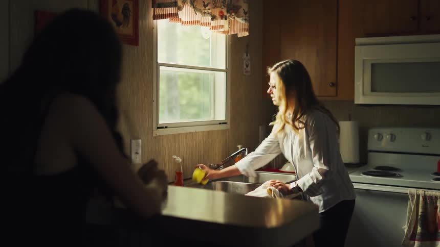 A woman's anger erupts as she screams at another person while washing dishes in a dramatically lit kitchen
