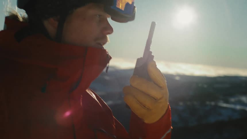 Close-up of a ski patrolman in a red jacket holding a walkie-talkie while on a snowy mountain with the sun flaring