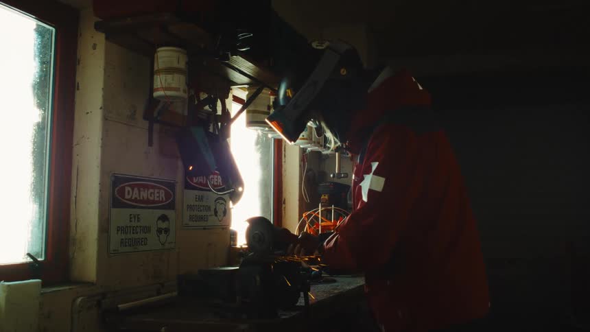 A worker grinds metal in a dark workshop, with sparks flying as the camera slowly pushes in on the action