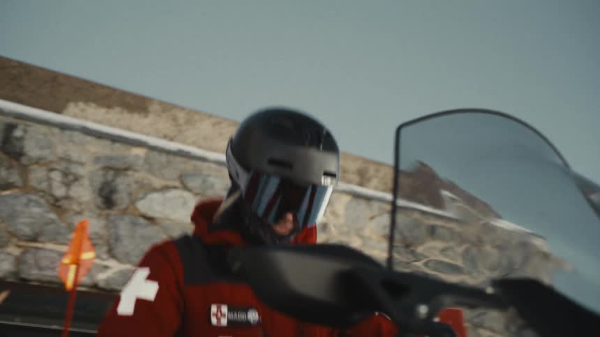 A Swiss ski patroller wearing a helmet and goggles starts up their snowmobile in the mountains