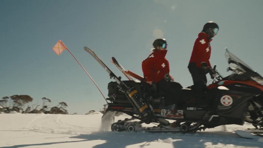 Two ski patrol members ride a snowmobile in slow motion across a sunny, snow-covered mountain landscape