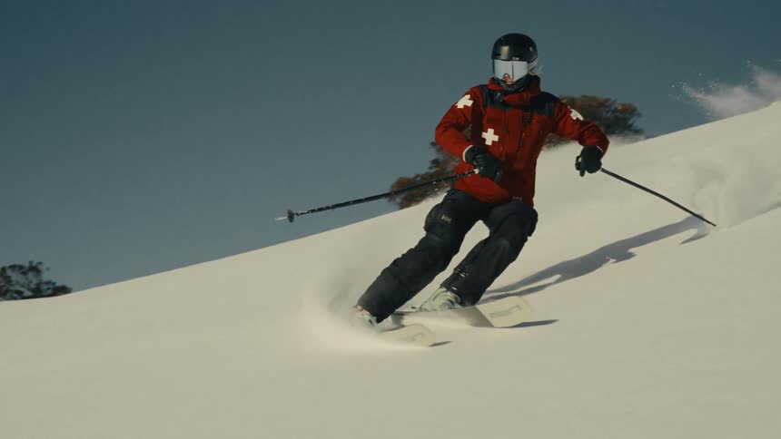 A professional ski patrol member skillfully carves through fresh powder, sending a massive wave of snow towards the camera in dramatic slow motion