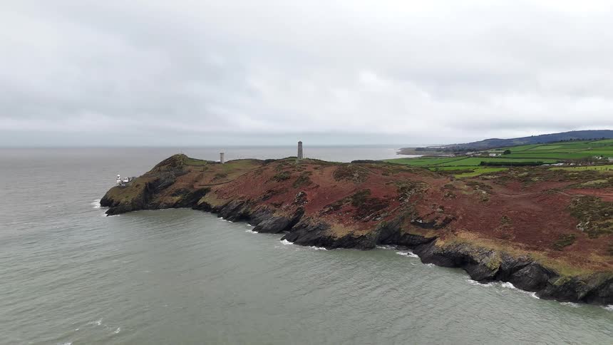 A wide aerial view of a rugged coastal headland with a lighthouse and a tower overlooking the sea under an overcast sky